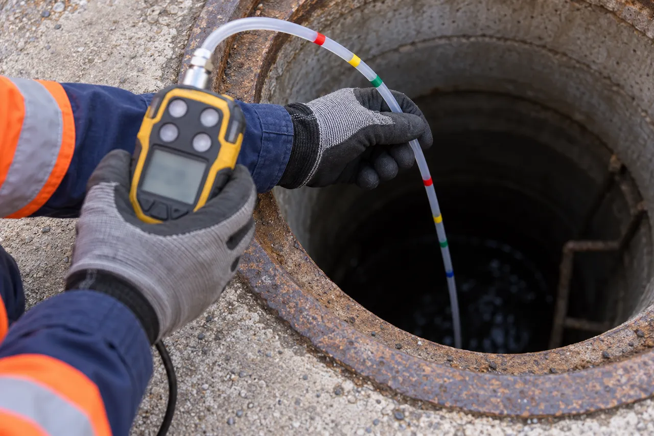 A gloved hand holding a portable gas detector while lowering a marked sampling hose into a manhole